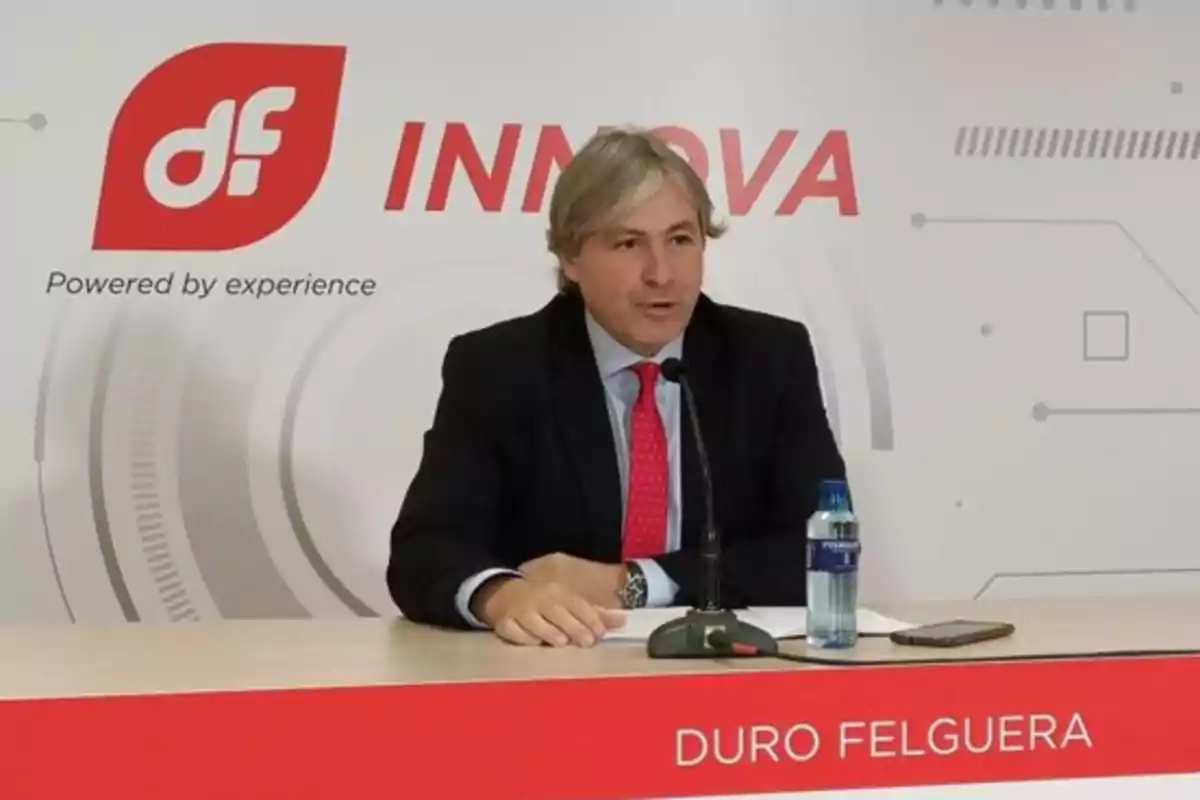 Un hombre en traje y corbata roja habla en una conferencia de prensa de Duro Felguera, con un micrófono frente a él y una botella de agua sobre la mesa.