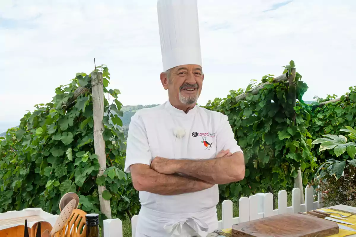 Un chef sonriente con uniforme blanco y gorro alto de cocina está de pie con los brazos cruzados frente a un fondo de viñedos y cielo despejado.