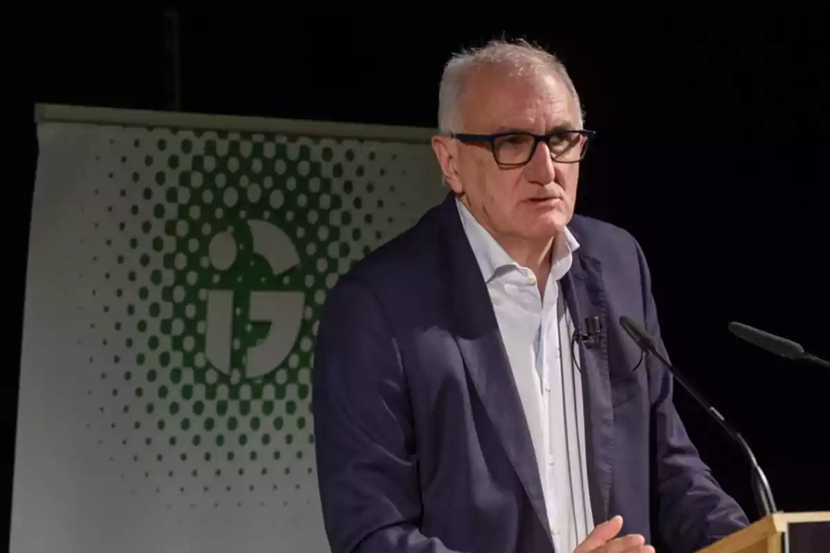 Un hombre con gafas y cabello canoso habla en un podio con un micrófono y un cartel con un diseño verde de fondo.