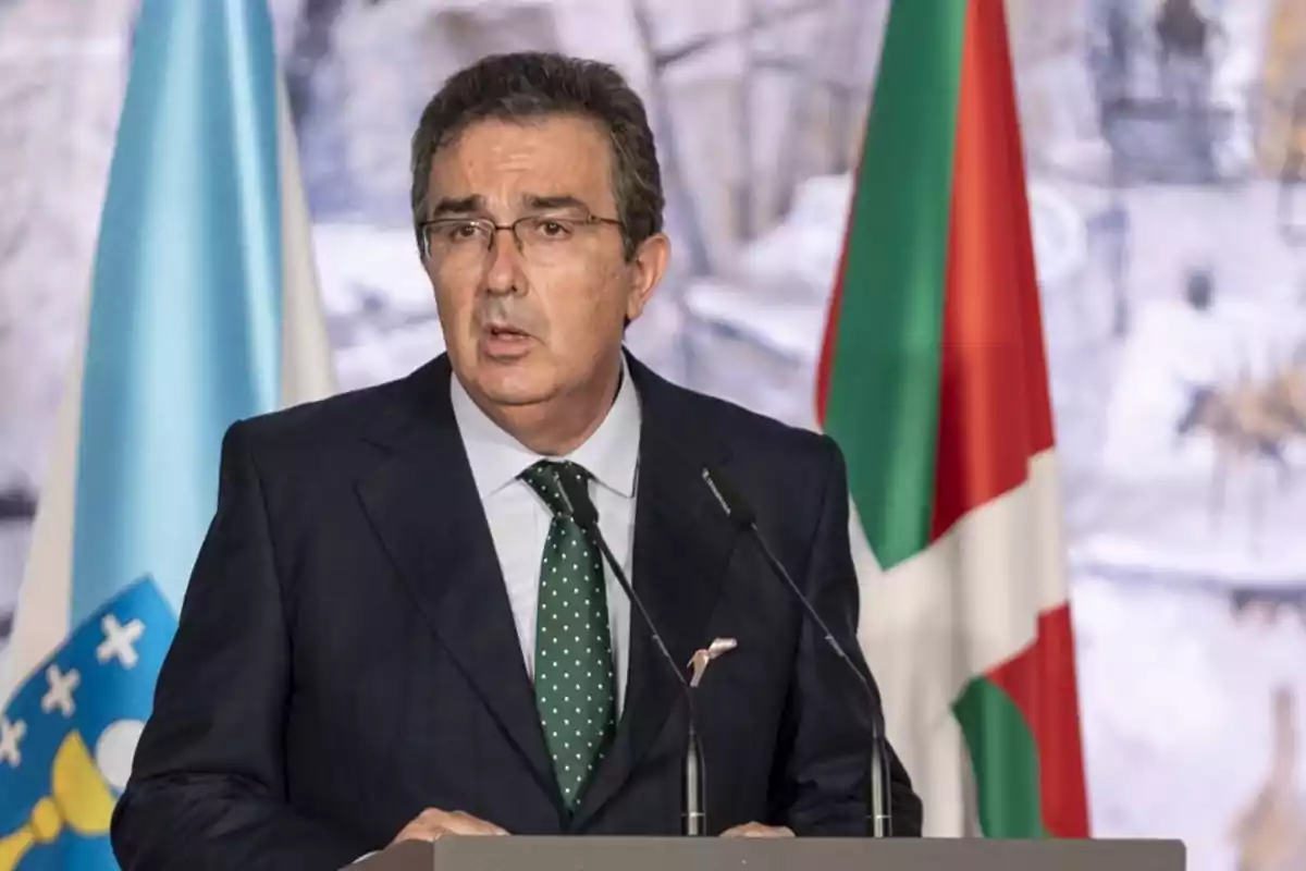 Hombre de traje oscuro y corbata verde con lunares hablando frente a dos micrófonos, con banderas de fondo Hombre de traje oscuro y corbata verde con lunares hablando frente a dos micrófonos, con banderas de fondo