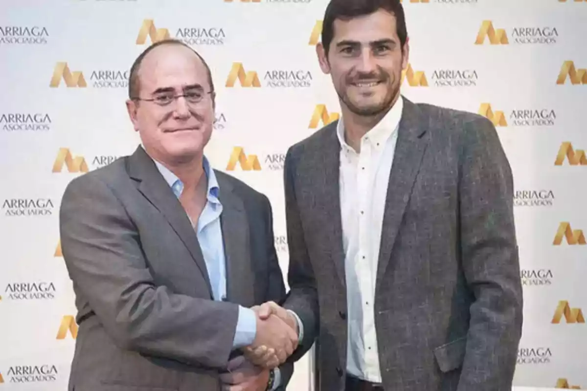 Dos hombres de traje estrechándose la mano frente a un fondo con el logo de Arriaga Asociados.