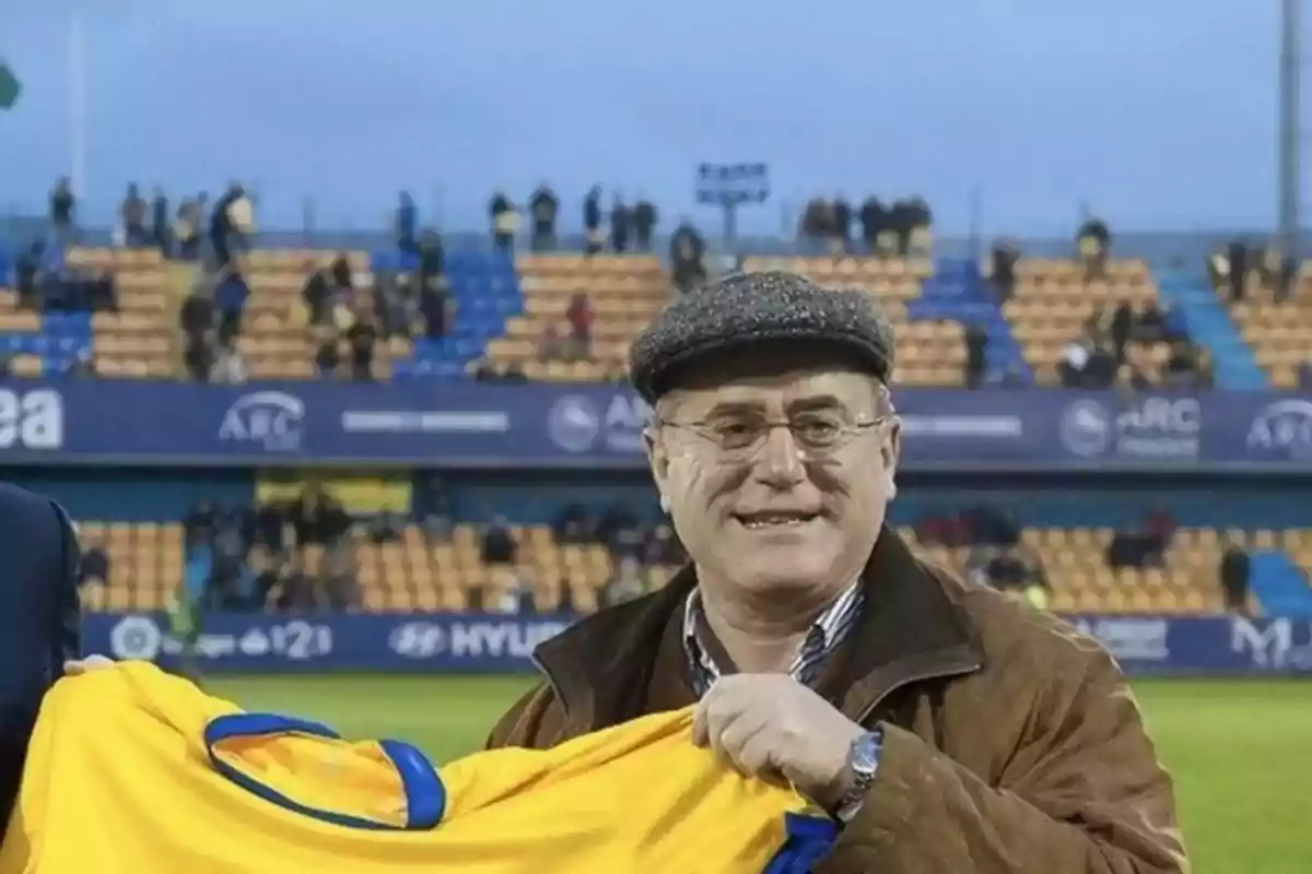 Un hombre sonriente con gafas y boina sostiene una camiseta amarilla en un estadio de fútbol.