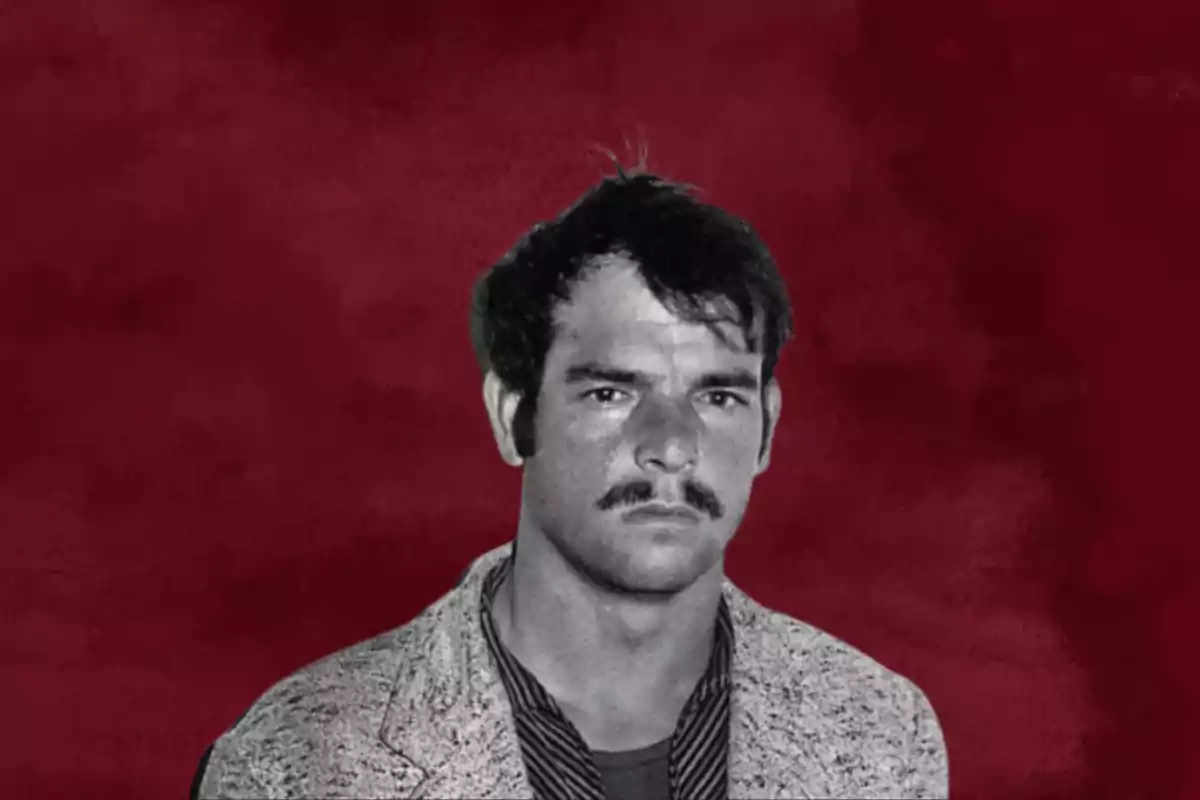 Un hombre con bigote y cabello oscuro sobre un fondo rojo.