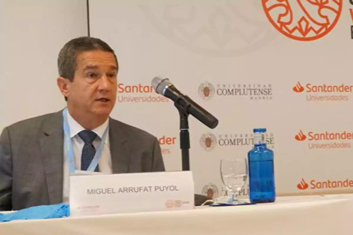 Hombre de traje hablando en una conferencia con un micrófono frente a él y una botella de agua sobre la mesa