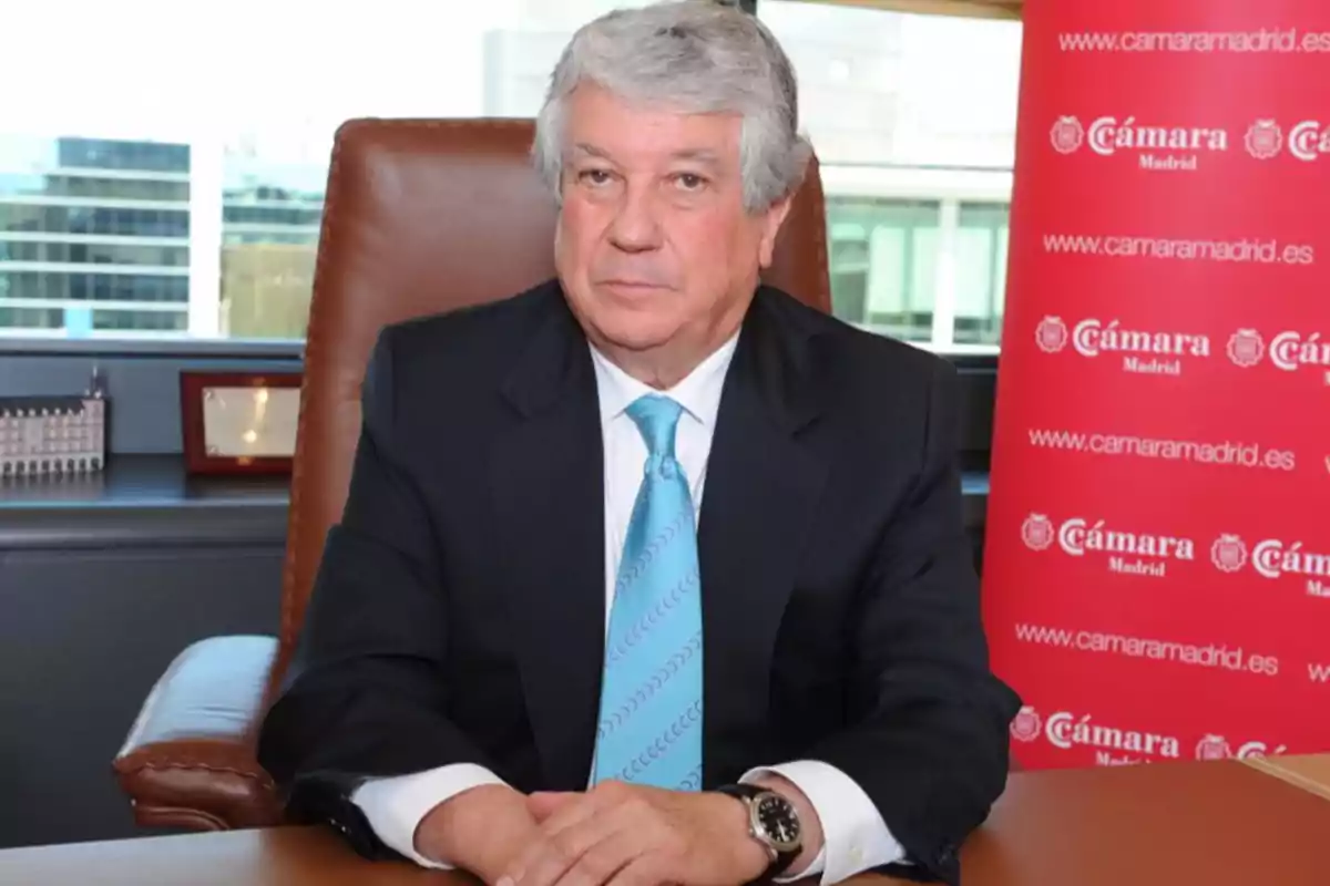 Un hombre de cabello canoso y traje oscuro está sentado en una oficina con un fondo que muestra un banner rojo de la Cámara de Madrid.