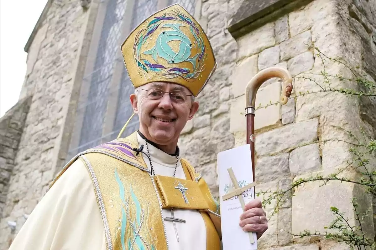 Un clérigo sonriente con vestimenta ceremonial dorada y mitra decorada sostiene un bastón y un documento frente a una iglesia de piedra.