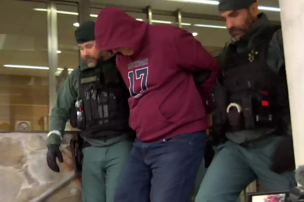 Un hombre con sudadera roja es escoltado por dos agentes de seguridad con uniformes verdes.