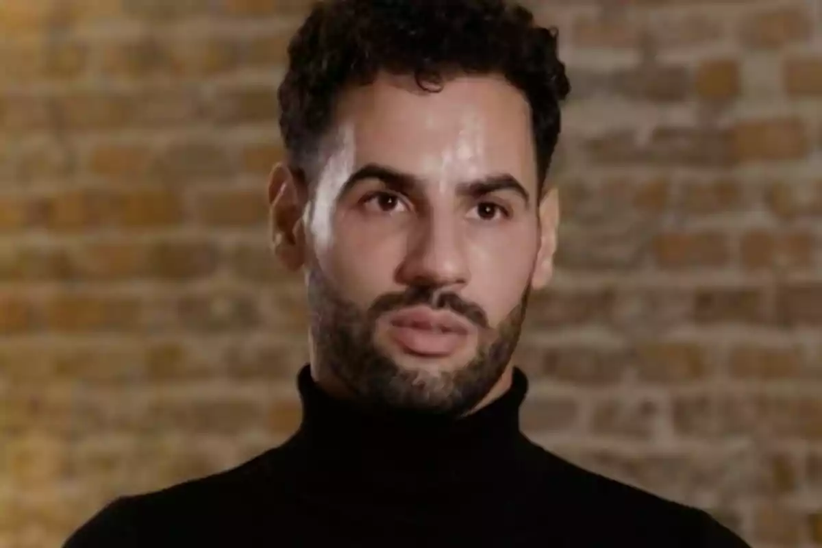 Asraf Beno con barba y cabello rizado lleva un suéter negro de cuello alto, con un fondo de pared de ladrillos en De Viernes.