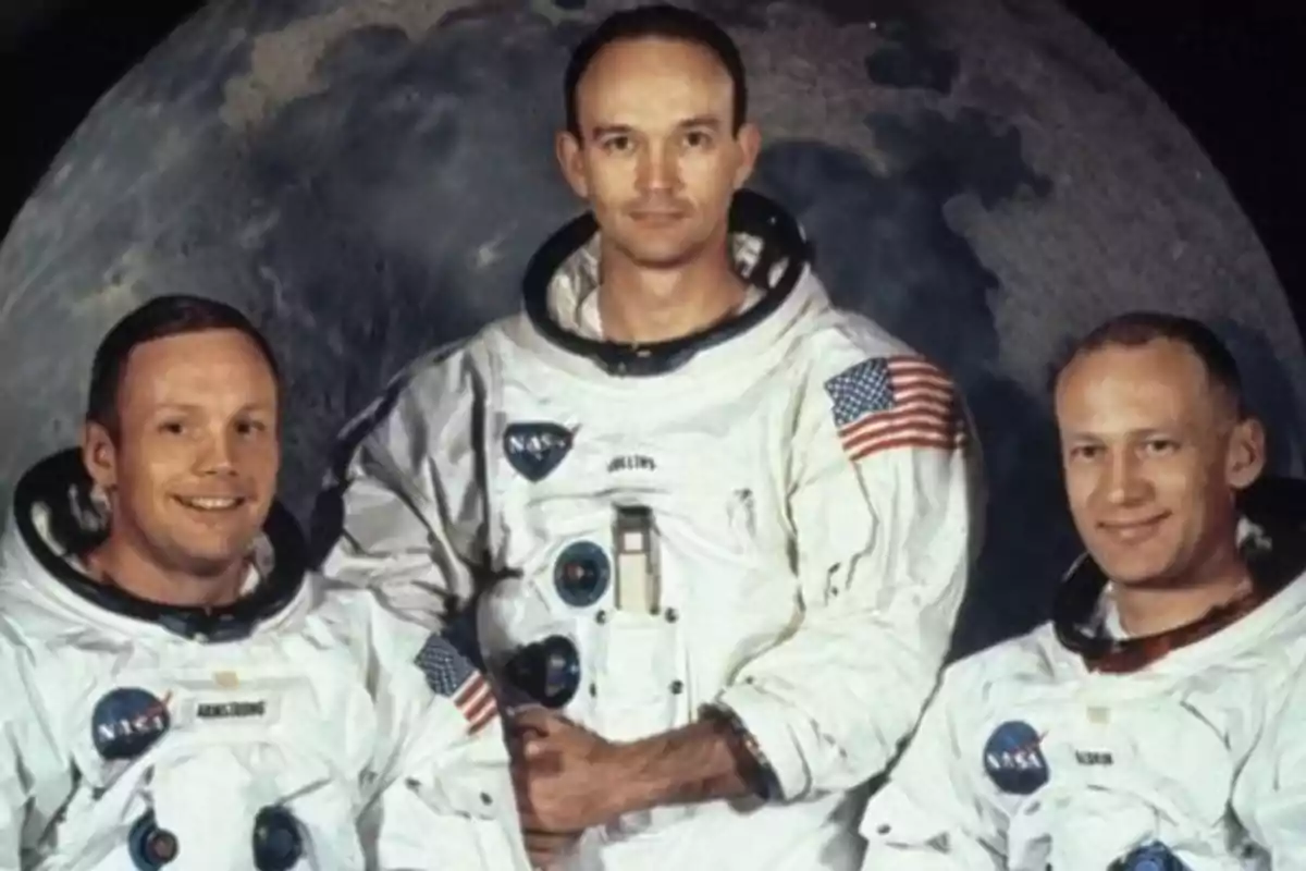 Tres astronautas con trajes espaciales de la NASA posan frente a una imagen de la luna.