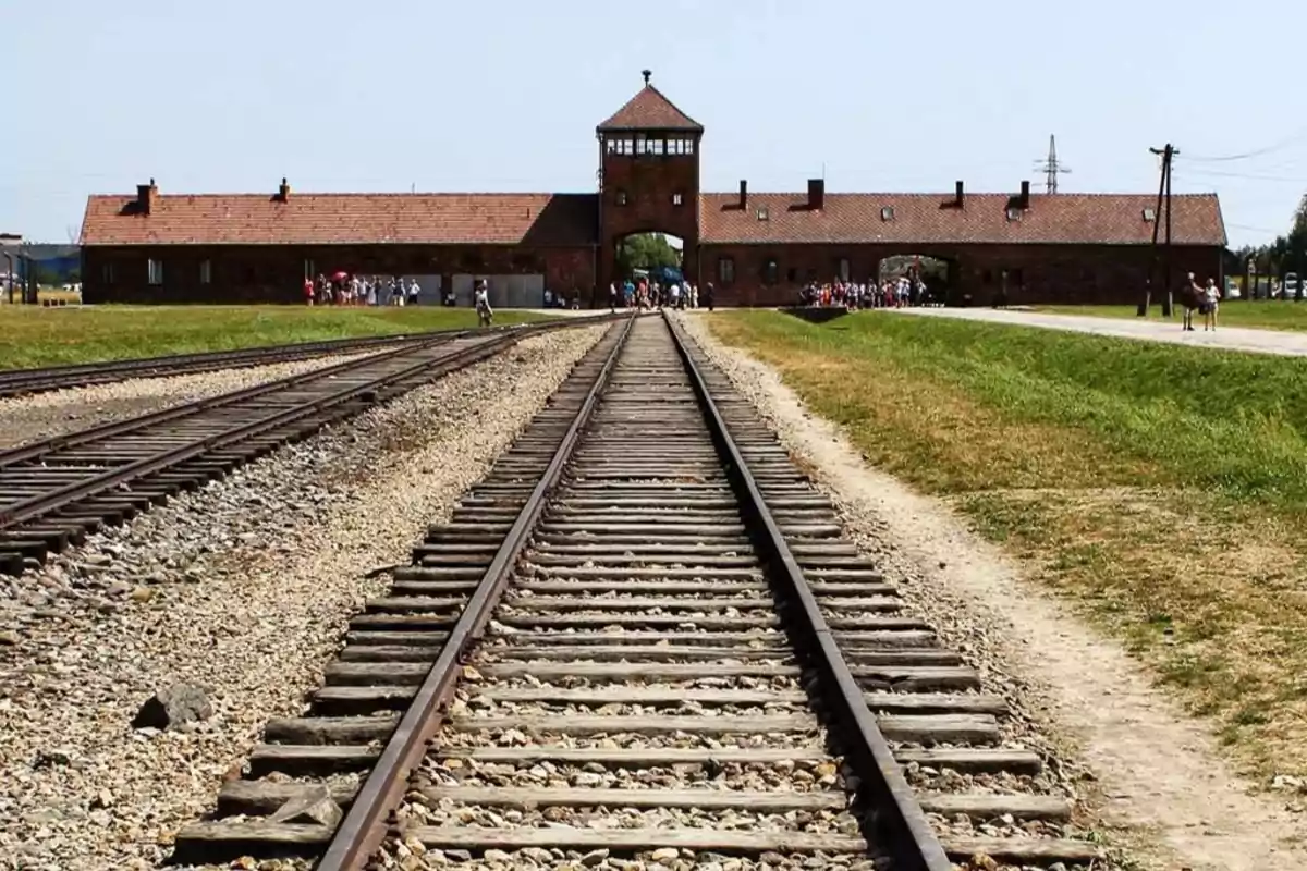 Vías de tren que conducen a la entrada de un edificio histórico de ladrillo con una torre central y un grupo de personas en el fondo.