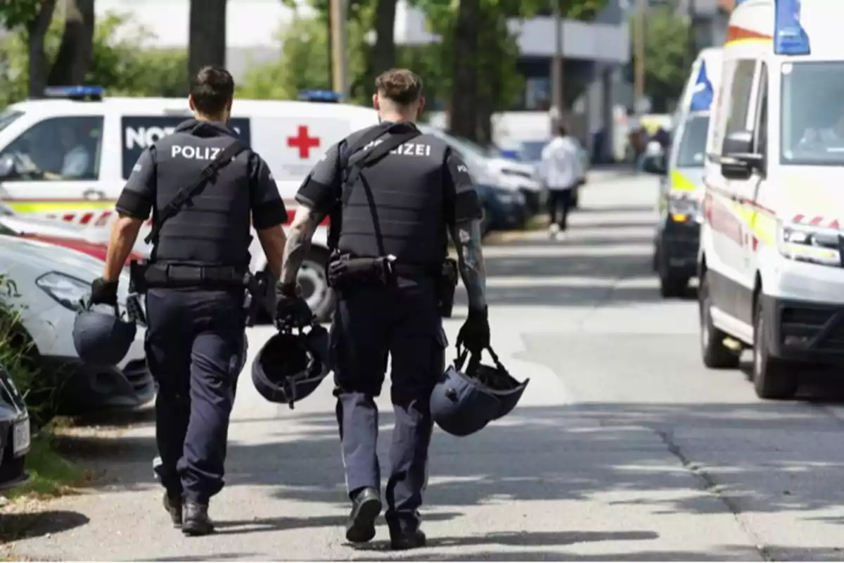 Dos policías de espaldas caminan por una calle llevando cascos en la mano mientras varias ambulancias y vehículos de emergencia están estacionados a su alrededor
