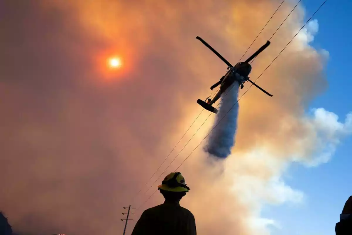 Un helicóptero arroja agua sobre un incendio mientras un bombero observa desde el suelo con el sol brillando a través del humo en el cielo en Los Ángeles. Un helicóptero arroja agua sobre un incendio mientras un bombero observa desde el suelo con el sol brillando a través del humo en el cielo en Los Ángeles.