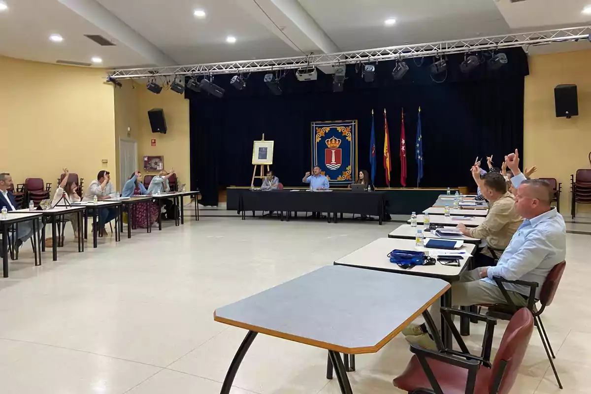 Una reunión de personas en una sala con mesas y sillas, algunas de ellas levantando la mano, con un escenario al fondo decorado con banderas y un escudo.