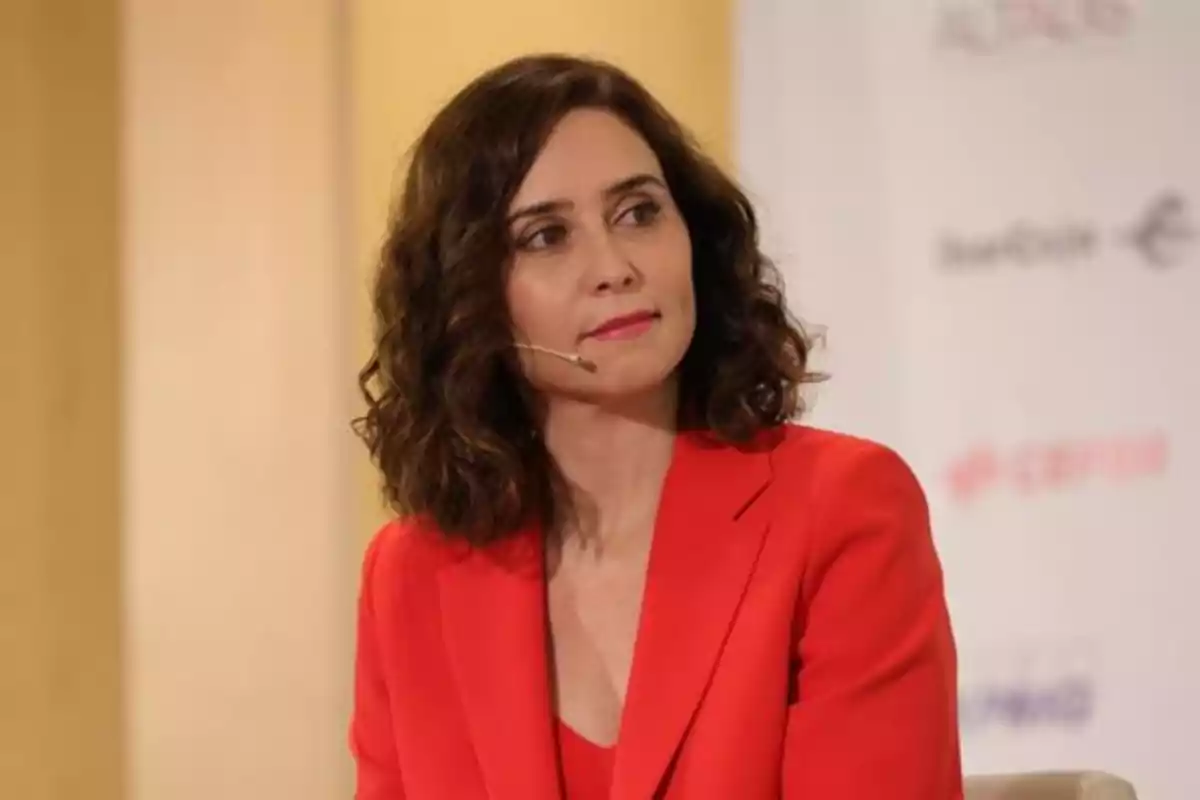 Mujer de cabello rizado y oscuro con chaqueta roja y micrófono de diadema sentada en un evento Mujer de cabello rizado y oscuro con chaqueta roja y micrófono de diadema sentada en un evento