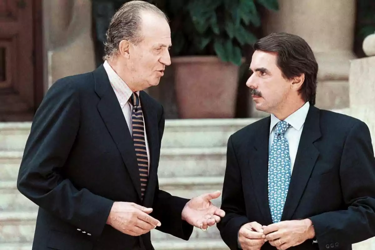 Dos hombres de traje conversando en un entorno formal con escaleras y plantas al fondo.