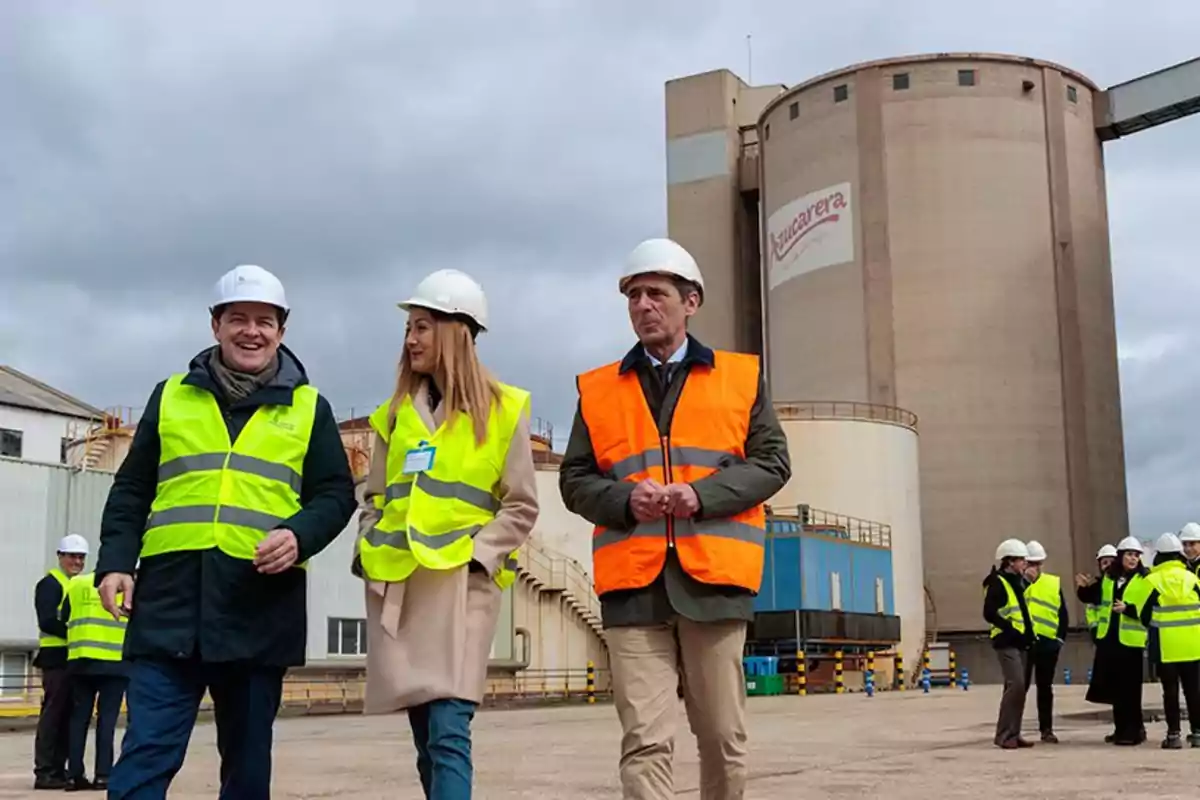 Personas con chalecos de seguridad y cascos blancos caminando frente a una planta industrial con un edificio cilíndrico grande al fondo. Personas con chalecos de seguridad y cascos blancos caminando frente a una planta industrial con un edificio cilíndrico grande al fondo.