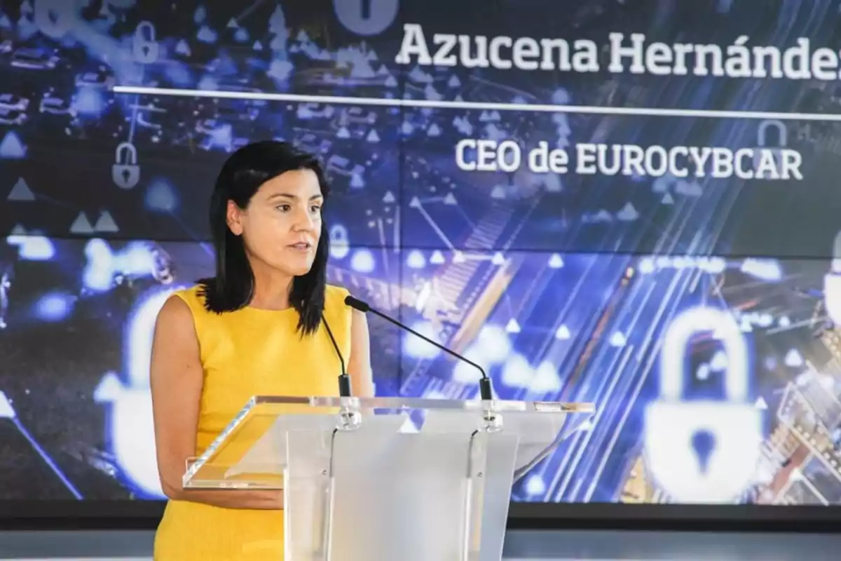 Una mujer con vestido amarillo hablando en un podio con un fondo digital que muestra su nombre y título como CEO de EUROCYBCAR.