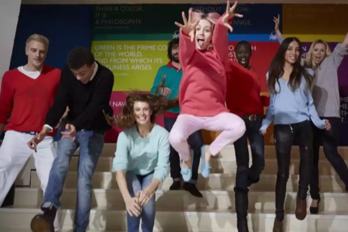 Un grupo de personas jóvenes saltando y sonriendo en una escalera, con un fondo colorido de carteles.