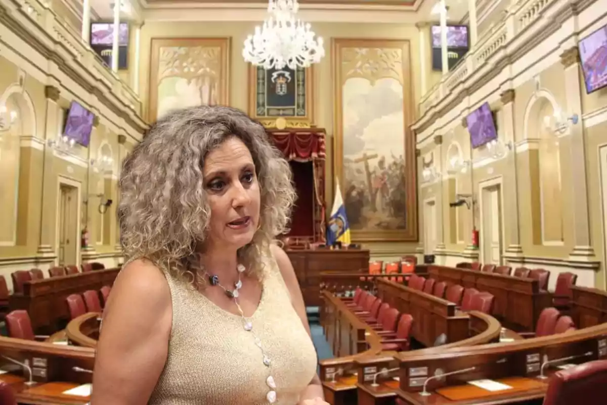 Una mujer de cabello rizado y canoso, vestida con una blusa sin mangas de color claro, se encuentra en el interior de una sala de reuniones o parlamento con asientos de madera y un gran candelabro colgando del techo.