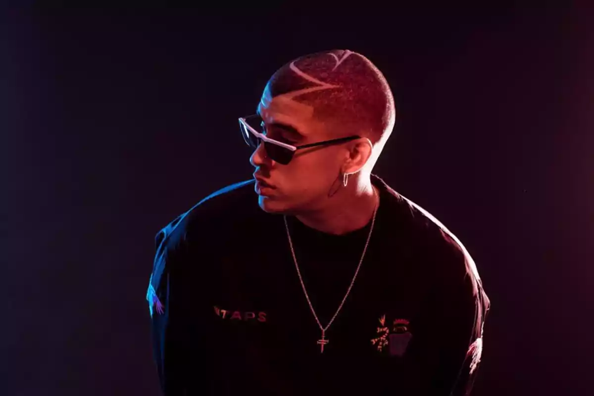 Un hombre con gafas de sol y un diseño en el cabello, vestido con una camiseta oscura y un collar, en un fondo oscuro iluminado con luces de colores. Un hombre con gafas de sol y un diseño en el cabello, vestido con una camiseta oscura y un collar, en un fondo oscuro iluminado con luces de colores.