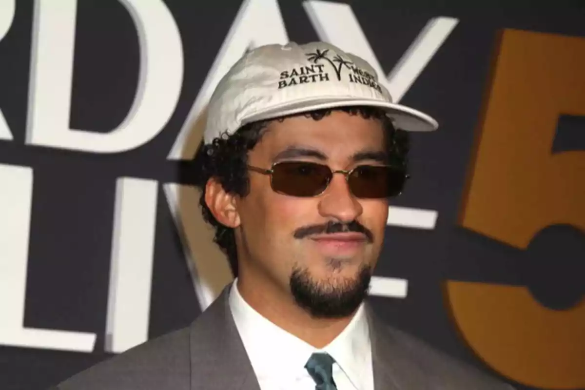 Un hombre con gafas de sol y gorra blanca está posando frente a un fondo con letras grandes.