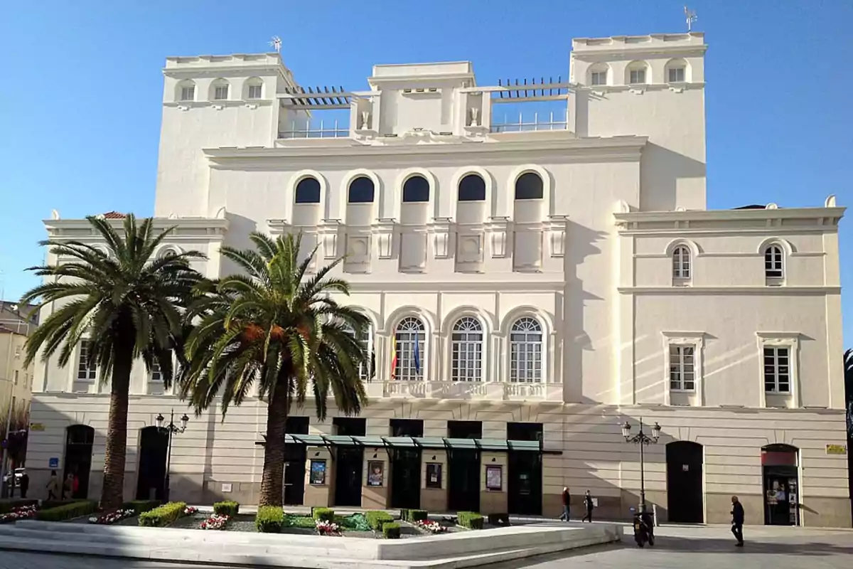 Edificio blanco de estilo neoclásico con palmeras en la entrada y cielo despejado.