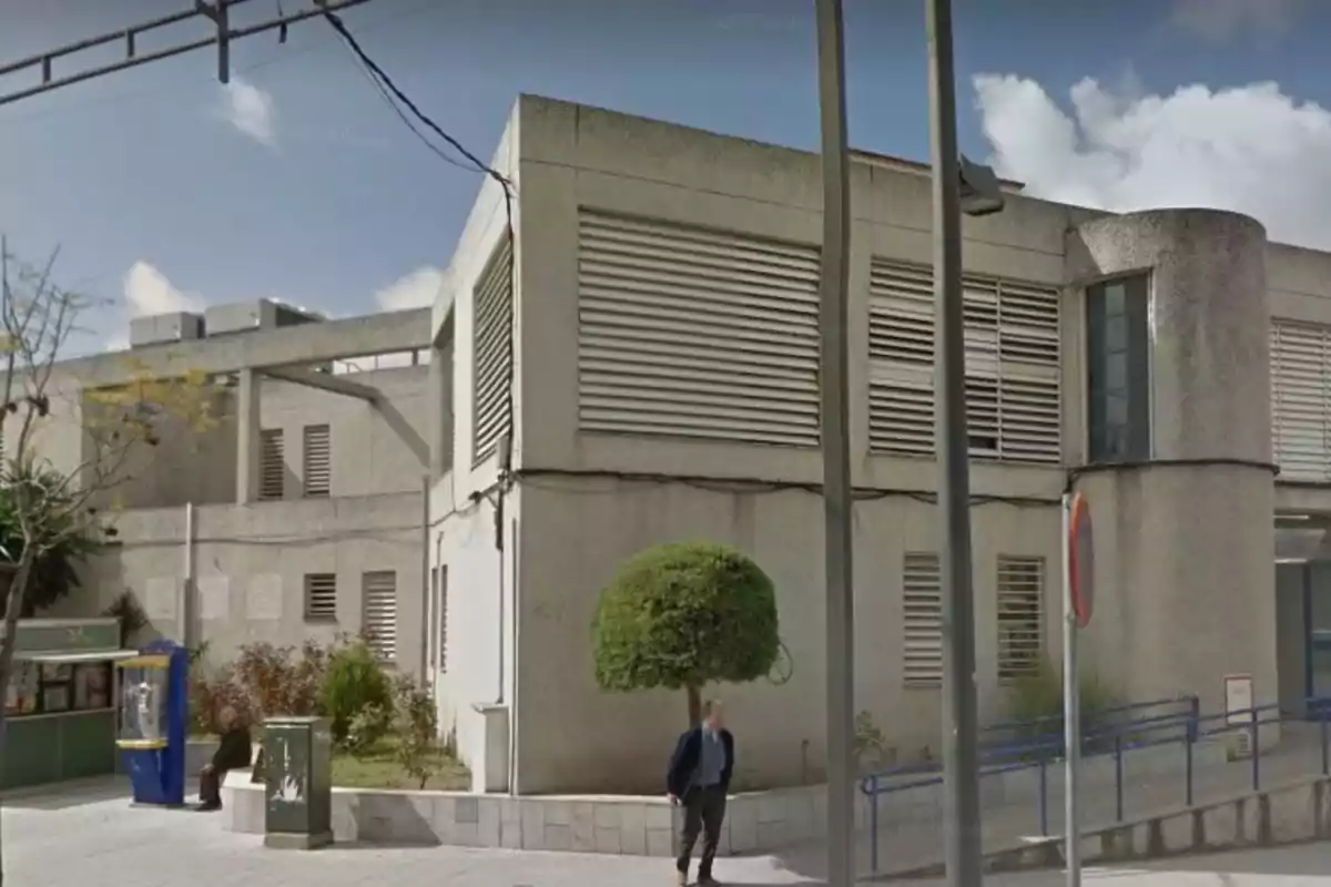 Edificio de concreto con ventanas de persianas y un pequeño jardín al frente, con una persona caminando y otra sentada cerca de una cabina telefónica.