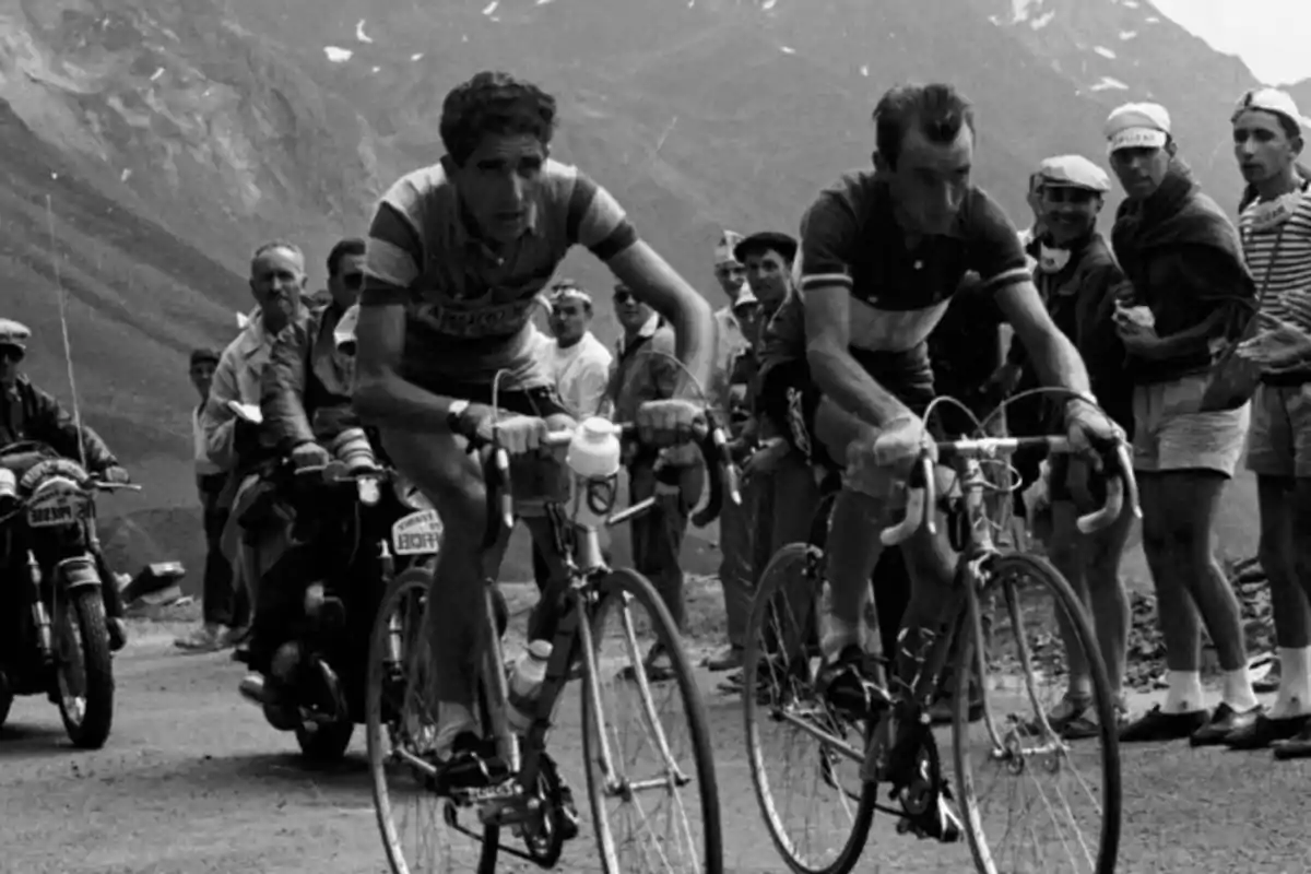 Dos ciclistas compiten en una carrera de montaña mientras son observados por espectadores y motociclistas en un paisaje montañoso.