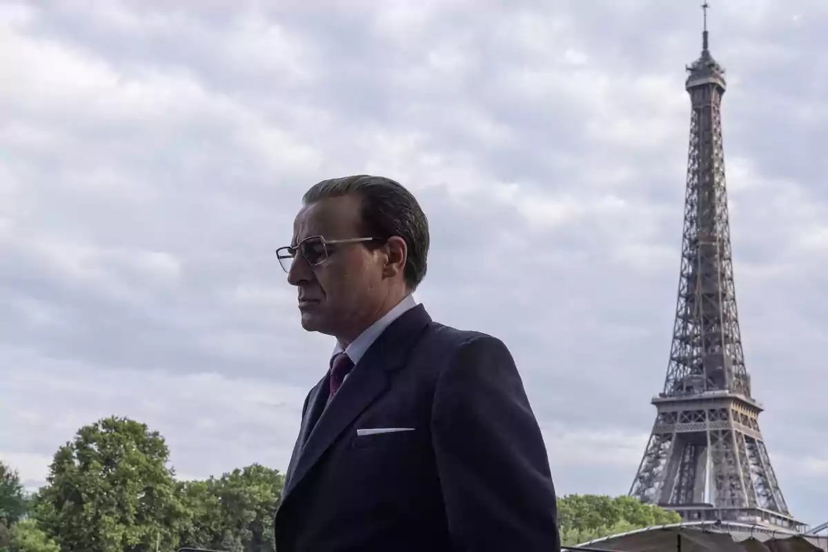 Hombre con gafas y traje oscuro de pie frente a la Torre Eiffel en un día nublado.