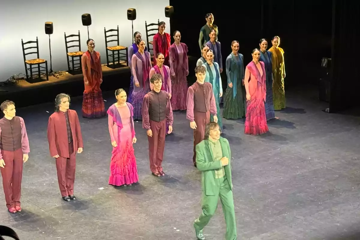 Un grupo de bailarines de flamenco en el escenario, vestidos con trajes coloridos, se encuentra en formación mientras un hombre con traje verde se adelanta.