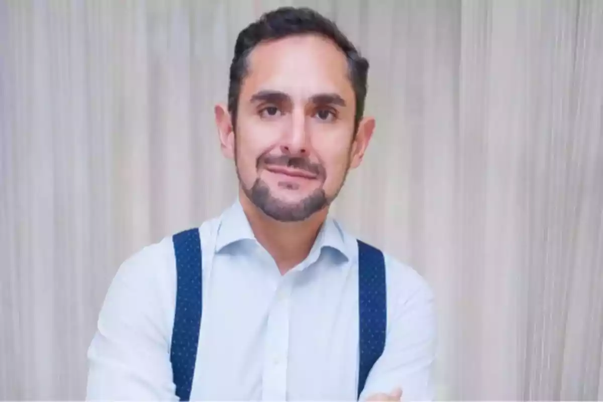 Hombre de cabello corto y barba, vestido con camisa clara y tirantes azules, posando con los brazos cruzados frente a un fondo claro. Hombre de cabello corto y barba, vestido con camisa clara y tirantes azules, posando con los brazos cruzados frente a un fondo claro.