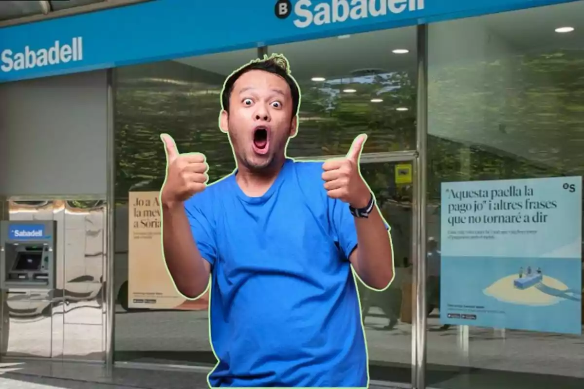 Un hombre con camiseta azul y expresión de sorpresa está frente a una sucursal del banco Sabadell, haciendo un gesto de aprobación con ambos pulgares hacia arriba.