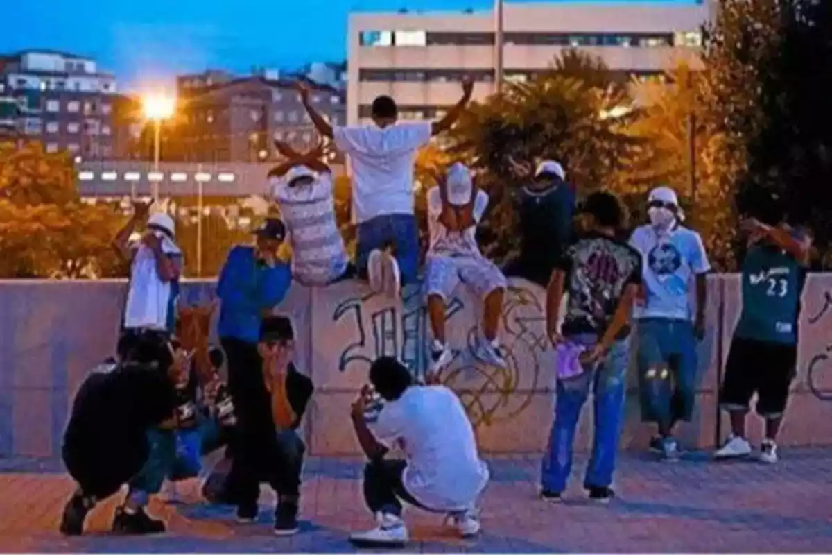 Un grupo de jóvenes posando en una plaza urbana al atardecer, algunos están sentados en un muro y otros agachados, con edificios y árboles al fondo.