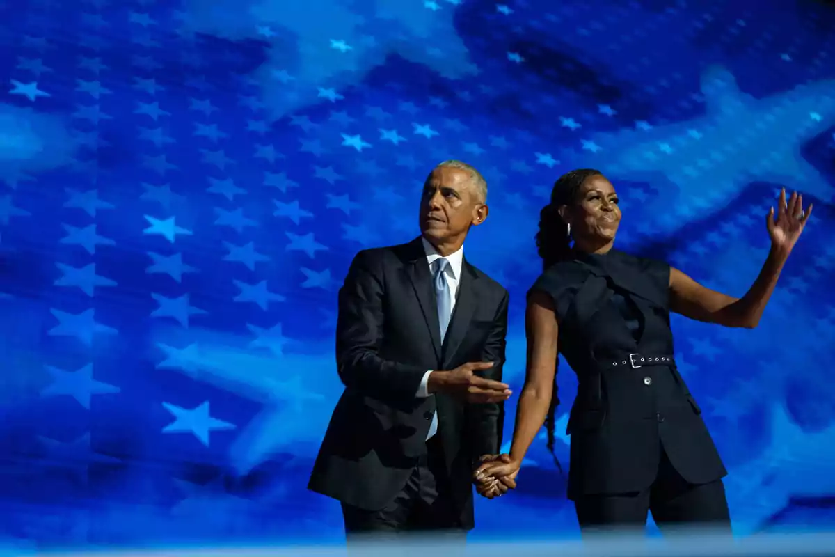 Barack y Michelle Obama de pie en un escenario con un fondo azul lleno de estrellas. Barack y Michelle Obama de pie en un escenario con un fondo azul lleno de estrellas.