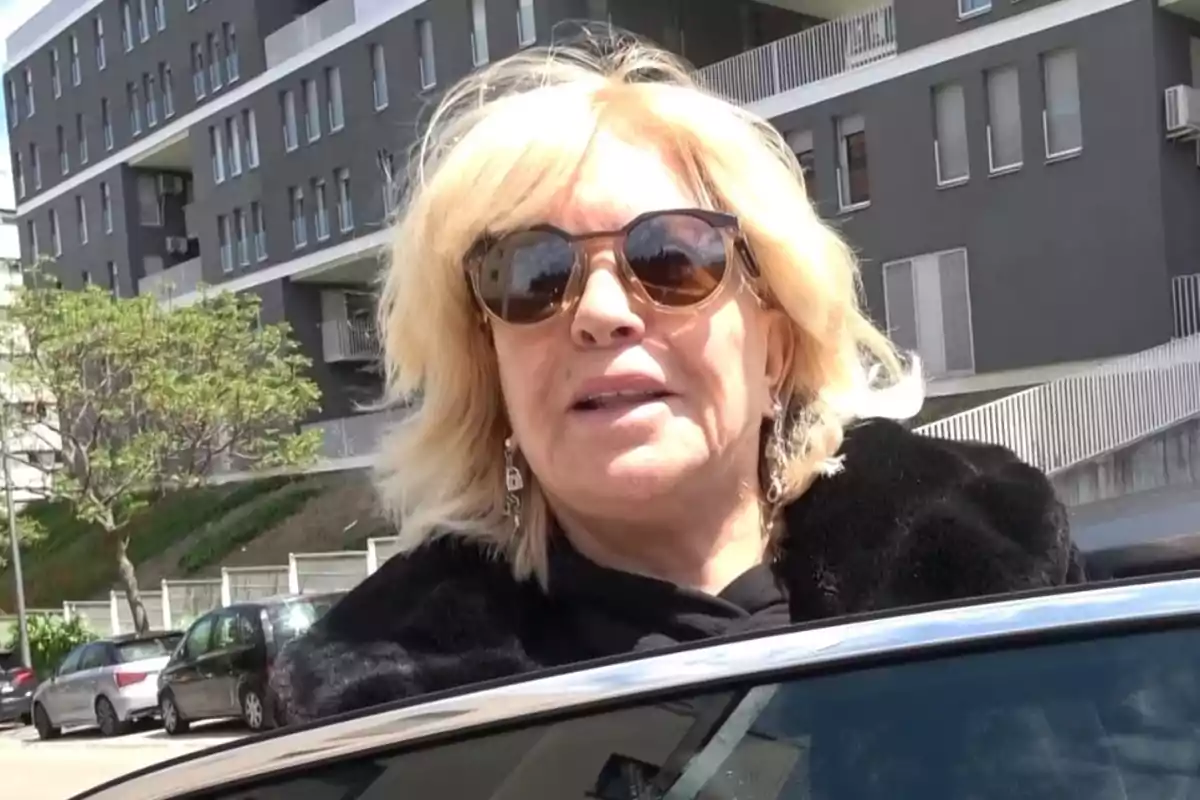 Bárbara Rey con gafas de sol y cabello rubio está de pie junto a un automóvil, con un edificio moderno de fondo. Bárbara Rey con gafas de sol y cabello rubio está de pie junto a un automóvil, con un edificio moderno de fondo.