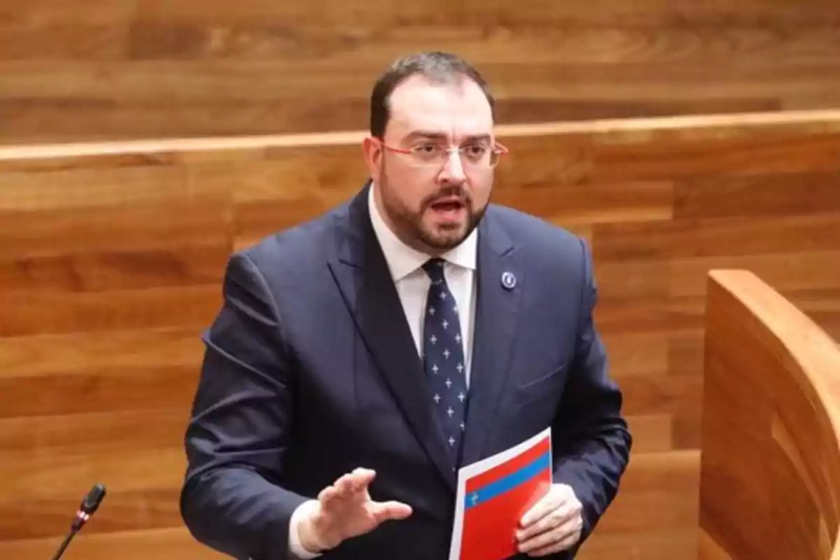 Hombre con barba y gafas hablando en un recinto de madera mientras sostiene una carpeta roja y azul