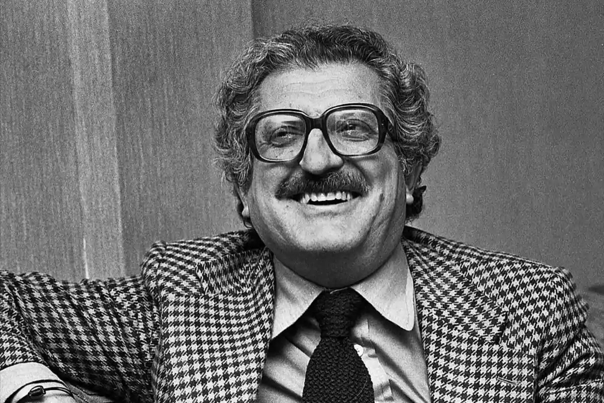 Un hombre con gafas grandes y bigote sonríe mientras lleva un traje de cuadros.