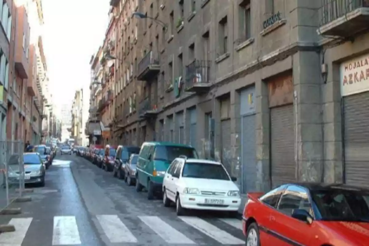 Calle estrecha con edificios altos a ambos lados y varios coches estacionados en la acera derecha.