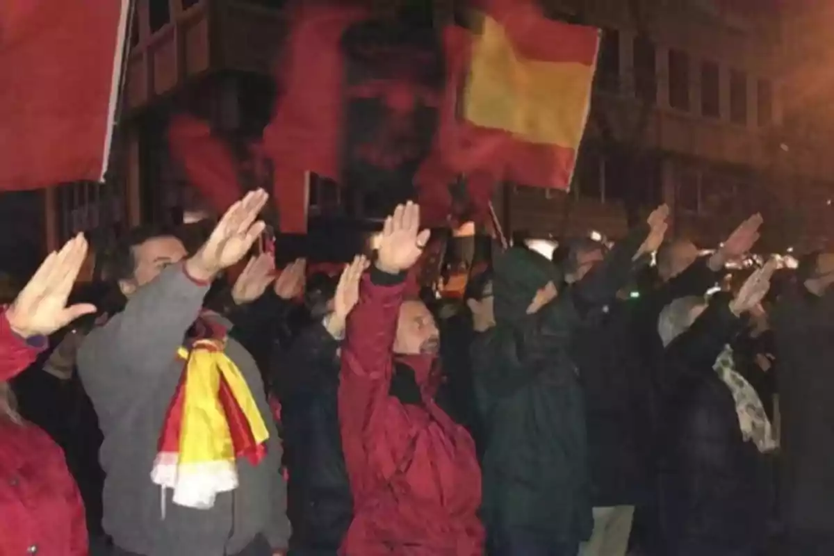 Un grupo de personas en una manifestación nocturna levantando el brazo derecho, con banderas rojas y amarillas ondeando en el fondo. Un grupo de personas en una manifestación nocturna levantando el brazo derecho, con banderas rojas y amarillas ondeando en el fondo.