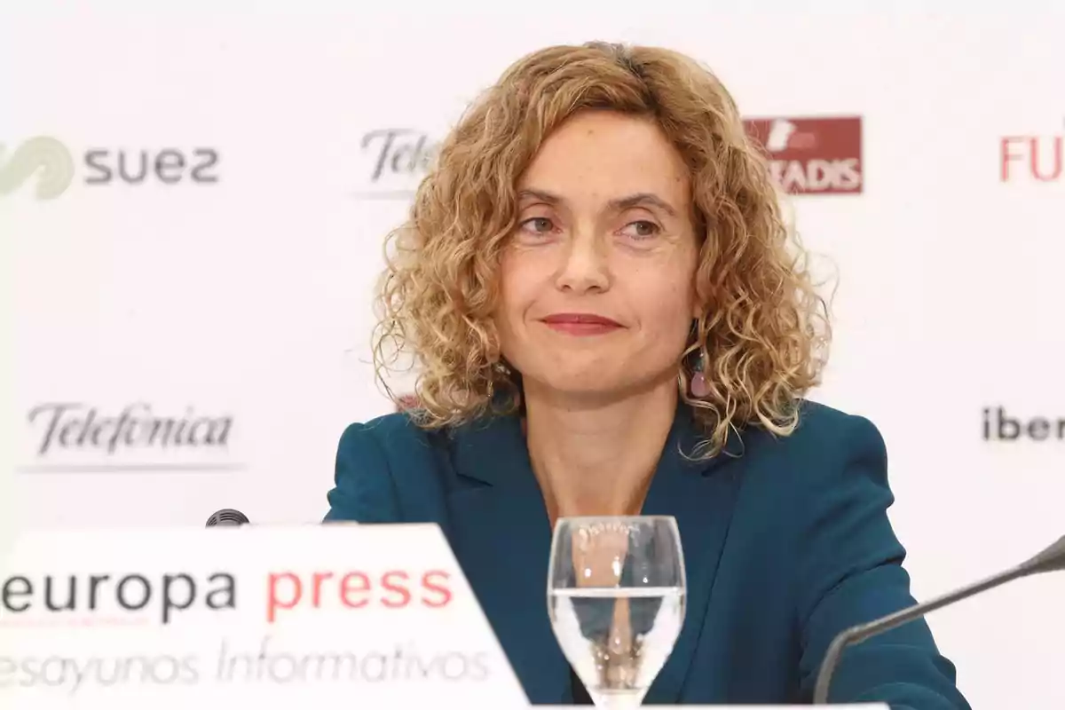 Una mujer con cabello rizado y rubio lleva un traje azul y está sentada en una mesa con un micrófono y un vaso de agua frente a ella, con un fondo que muestra logotipos de empresas. Una mujer con cabello rizado y rubio lleva un traje azul y está sentada en una mesa con un micrófono y un vaso de agua frente a ella, con un fondo que muestra logotipos de empresas.