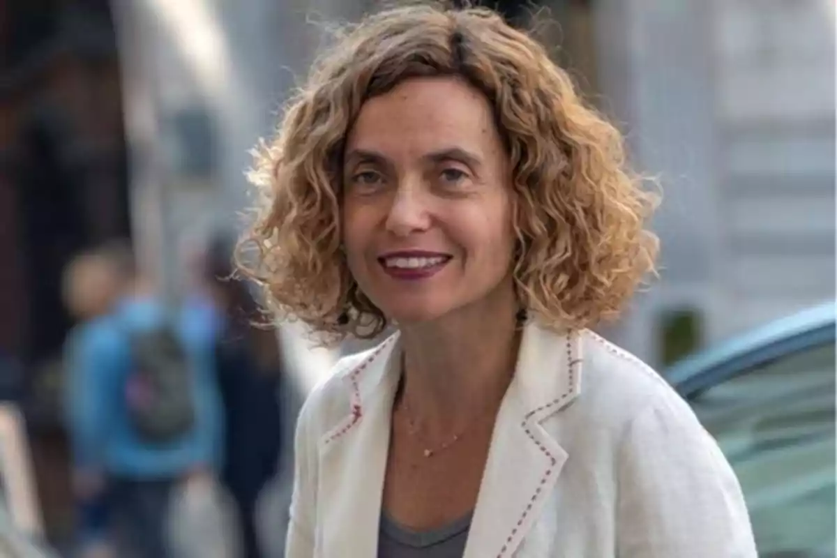 Una mujer con cabello rizado y rubio, vestida con una chaqueta blanca, sonriendo mientras está de pie en una calle urbana.