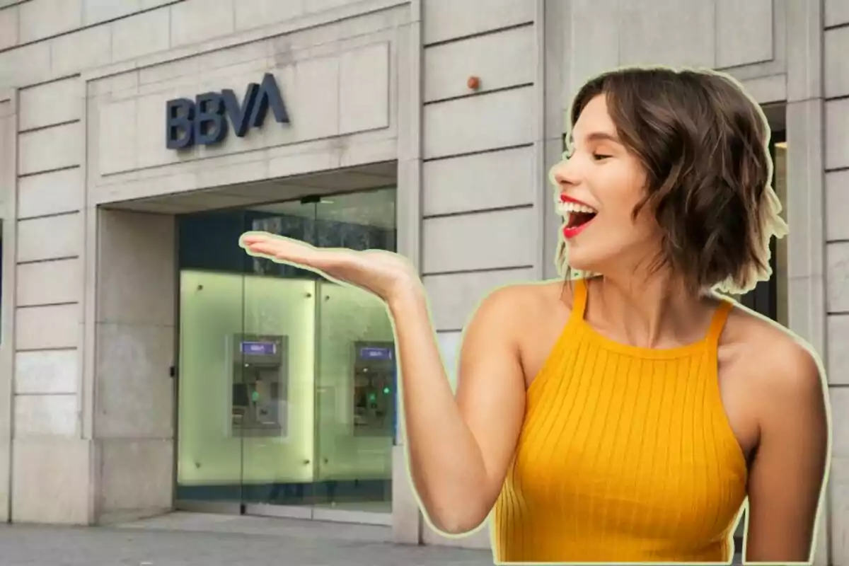 Una mujer sonriente con un vestido amarillo sostiene su mano extendida frente a una sucursal del banco BBVA.