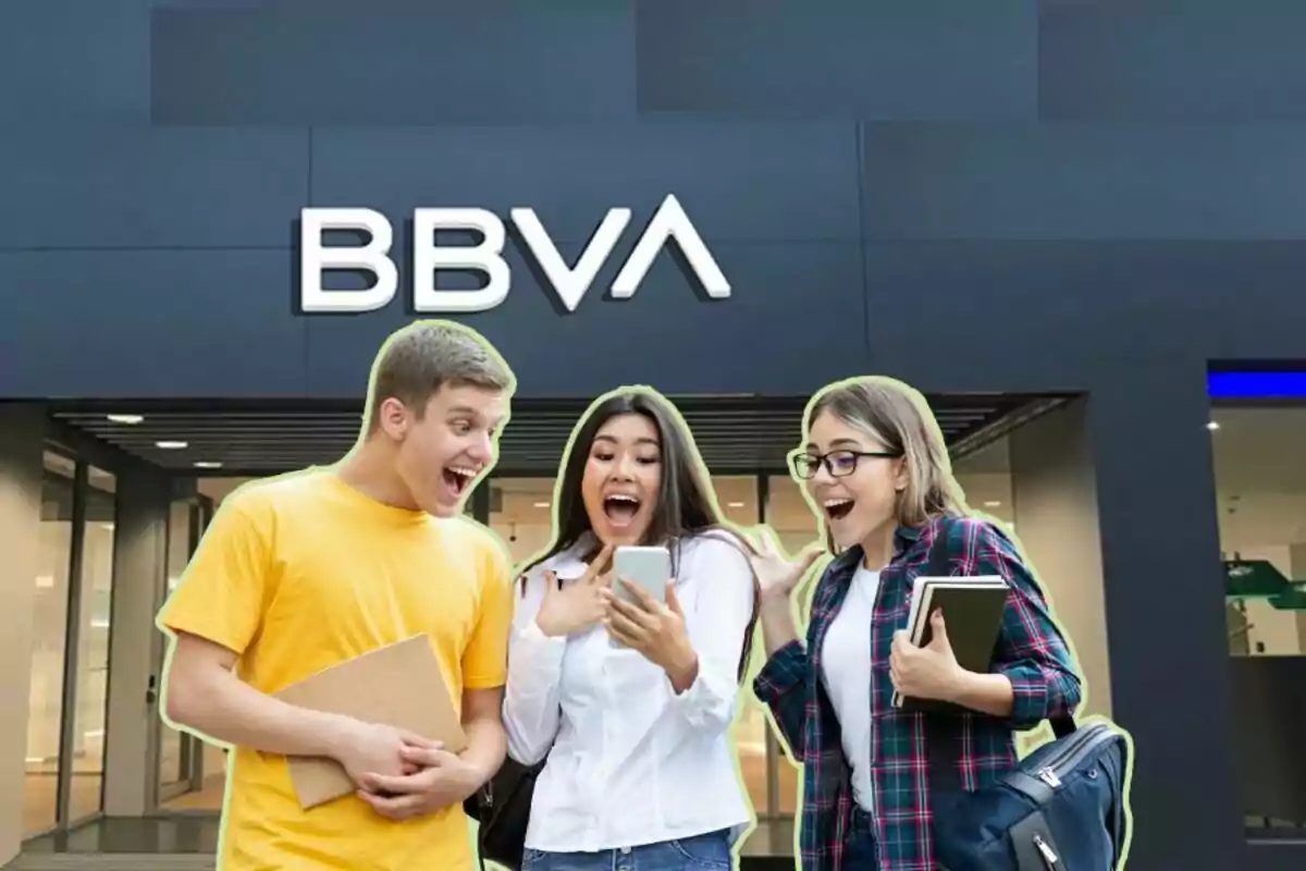 Tres personas jóvenes emocionadas mirando un teléfono móvil frente a una sucursal del banco BBVA.