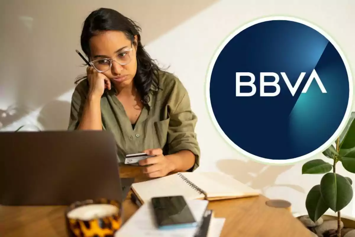 Mujer pensativa con gafas frente a una computadora portátil sosteniendo una tarjeta de crédito, con el logo de BBVA al lado.