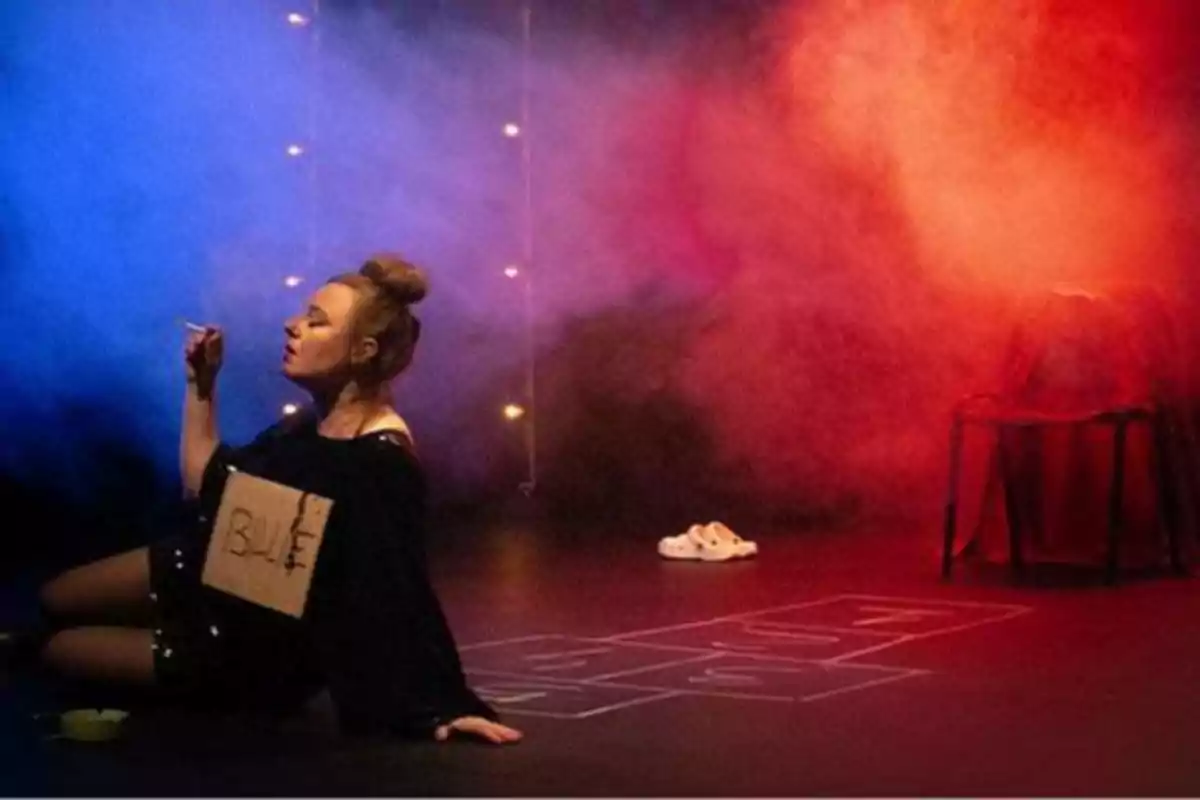 Una mujer sentada en el suelo de un escenario con luces de colores y humo, sosteniendo un cigarrillo y con un cartel que dice 