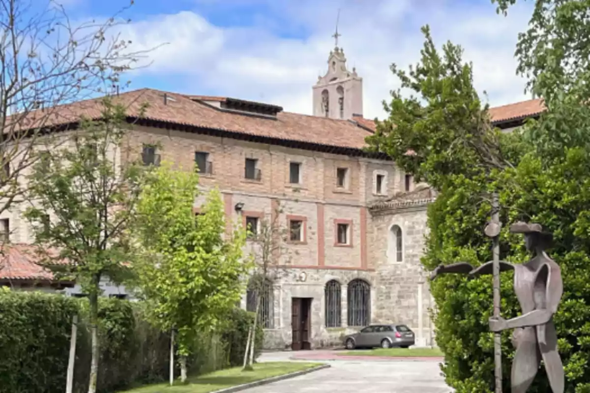 Edificio histórico de piedra con techo de tejas rojas, rodeado de árboles y vegetación, con una estatua de metal en primer plano y un coche estacionado frente a la entrada.