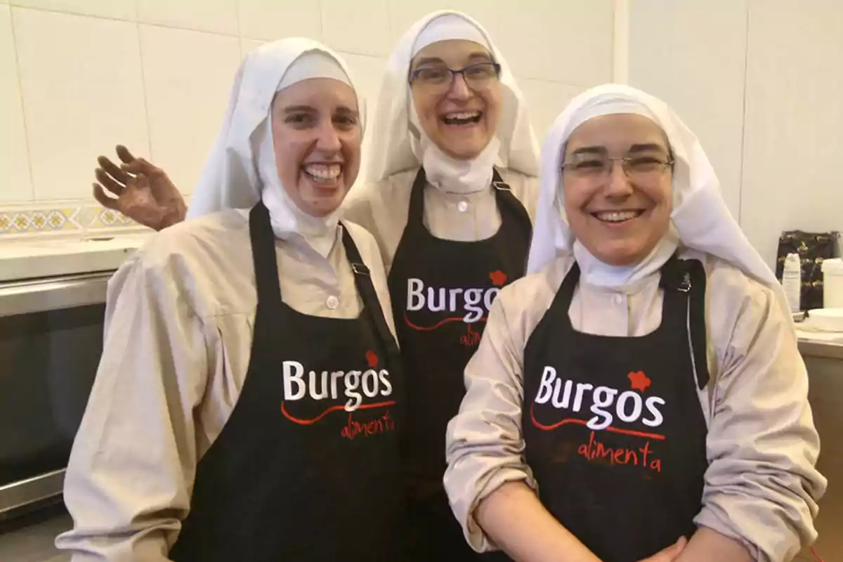 Tres monjas sonrientes con delantales negros que dicen "Burgos alimenta" en una cocina.