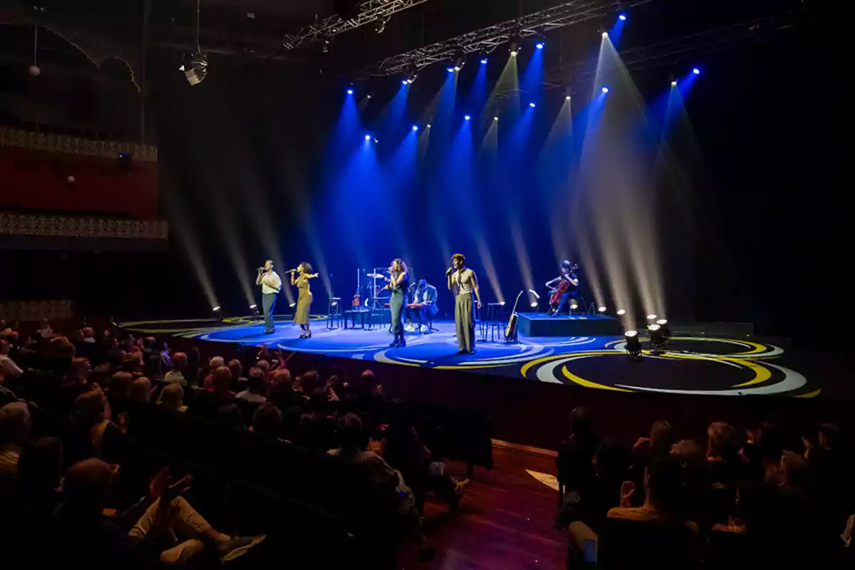 Un grupo de músicos y cantantes se presenta en un escenario iluminado con luces azules y blancas frente a una audiencia en un teatro.