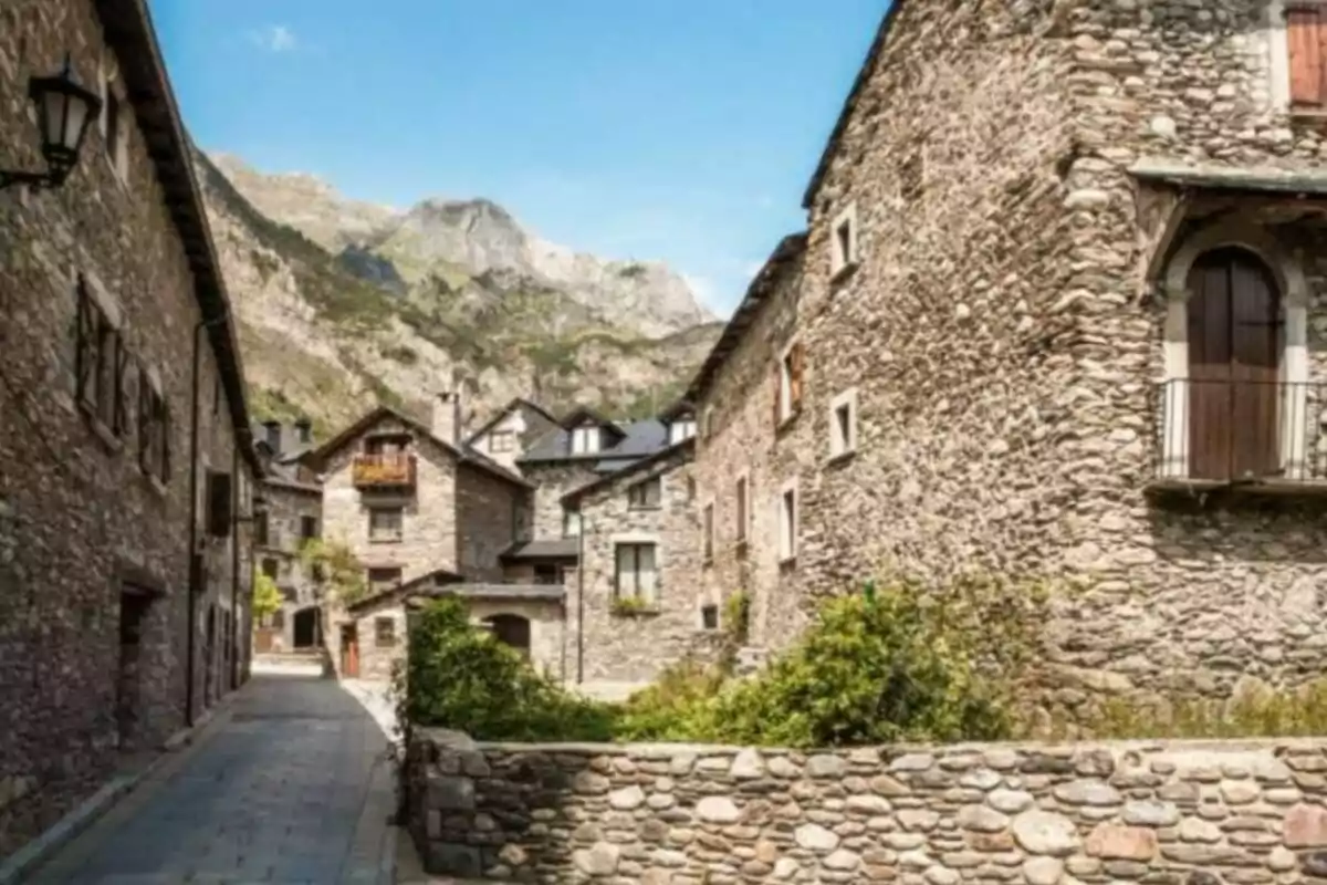 Calle estrecha de un pueblo con casas de piedra y montañas al fondo bajo un cielo despejado.