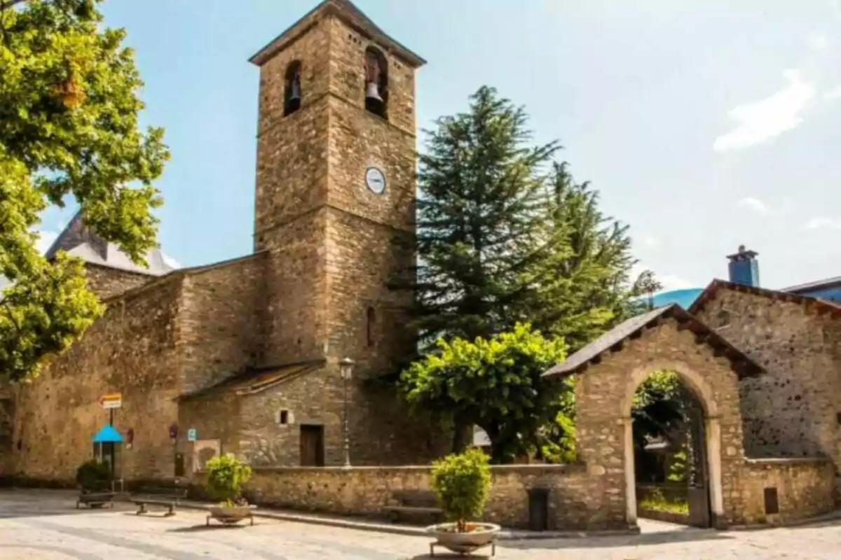 Torre de piedra de una iglesia con reloj en un entorno arbolado y soleado.